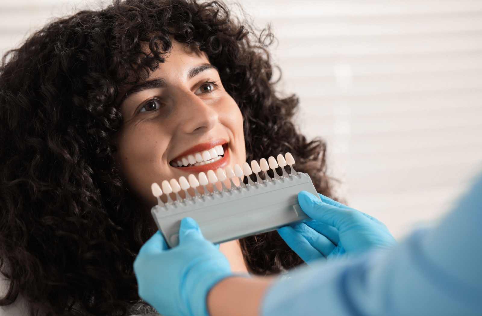 Person smiling during a dental consultation while a professional compares veneer shades using a sample palette in a bright room